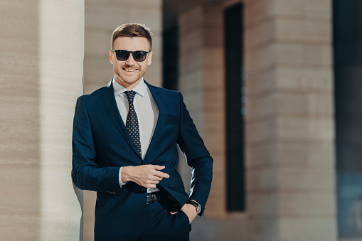 Positive glad young male wears formal suit, wears trendy shades, keeps hand in pocket, stands against office interior, being in good mood after successful business meeting. Leadership concept