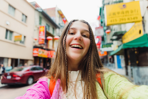 Self-portrait of smiling female traveler with long hair in colourful sweater doing blog in San Francisco Chinatown district, the United States