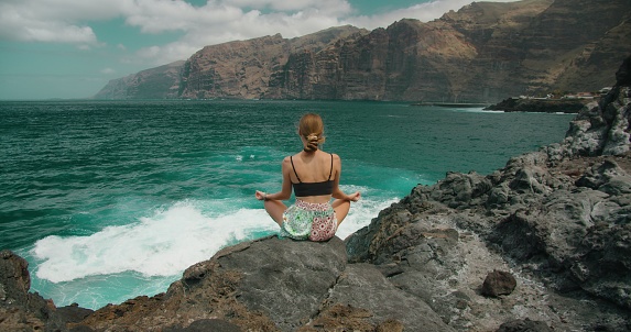 Back view of woman sitting in lotus pose and looking at tranquil sea surface, high mountain. Lady travels in Tenerife. Meditatation, relaxing exercises.