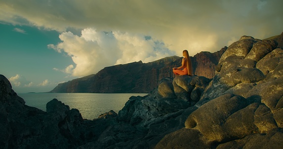 Woman practices yoga and meditates in lotus position on ocean rocky beach. Rear view of beautiful long haired adult girl silhouette on sea at sunset.
