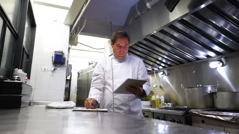 Chef checking the ingredients of a menu on tablet and checking the inventory on a document at a restaurant