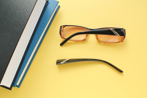 Old broken eyeglasses with damaged legs and books on yellow background. Poor eyesight. Repair concept. Idea of health. Failure optic eyewear. Breakage of vision correction glasses. Close up, flat lay