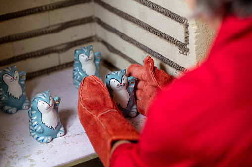 Mature craftswoman taking clay products from pottery kiln, handwork in pottery workshop.