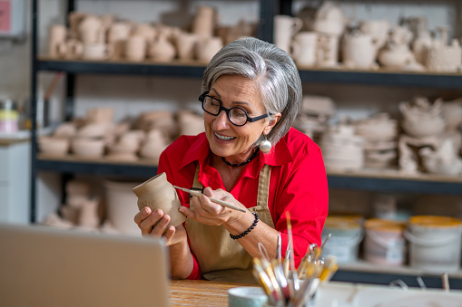 Mature woman painting pottery earthenware product in front of laptop while live streaming.