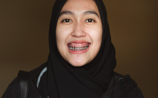 A close-up of a smiling young Asian Muslim woman with braces, smiling confidently at the camera