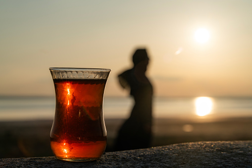 tea in glass cup and sunset view.standing woman silhouette.  Slender teacup. The sun is setting over the lake. Shot with a full-frame camera.