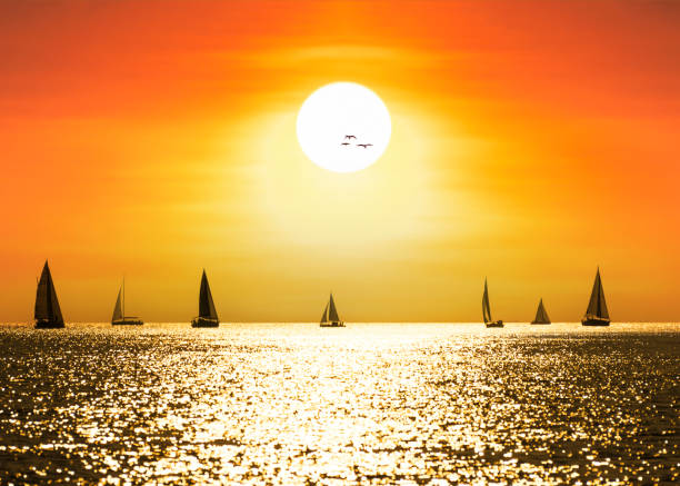 Sun shinning through fluffy clouds on a windy day while sailing boats are racing at the horizon. Dark blue wavy sea in front.