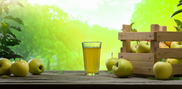 Apple harvest time. Glass of apple juice and ripe yellow apples in a crate on wooden table. Golden apples on the garden table