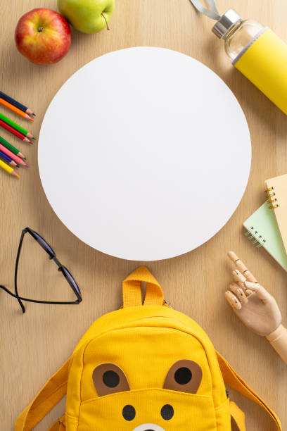 istock Playgroup concept. Vertical top view of crayons, copybooks, glasses, mannequin hand, backpack in shape of cute animal, water bottle, apples on wooden table backdrop with circle for text or advert 1534524095