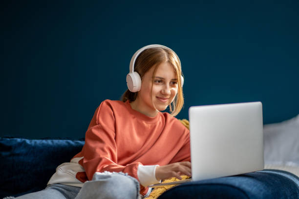 улыбающаяся девочка-подросток в наушниках печатает на ноутбуке - sofa music teenager friendship стоковые фото и изображения