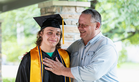 A teenage boy graduating from high school, wearing his cap and gown, poses for a portrait with his father. The graduate is smiling at the camera and dad is looking at him with pride, arm around his shoulder and hand on his chest.