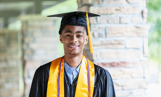 Headshot of a young African-American man graduating from high school. He is standing outside his school in cap and gown, smiling at the camera.