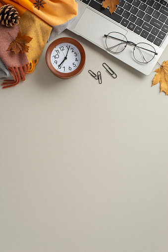 Autumn-infused work environment. Overhead vertical shot of laptop, glasses, cozy checkered blanket, golden maple leaves, natural pine cone, alarm clock, soft grey backdrop with space for text or promo