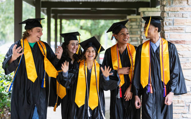 A group of five multiracial high school graduates standing together outside the school, wearing their caps and gowns, smiling.