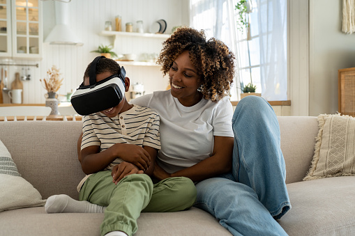 Happy joyful African American family mother and son using VR headset while relaxing together on sofa, laughing mom and child having fun while playing virtual reality games at home