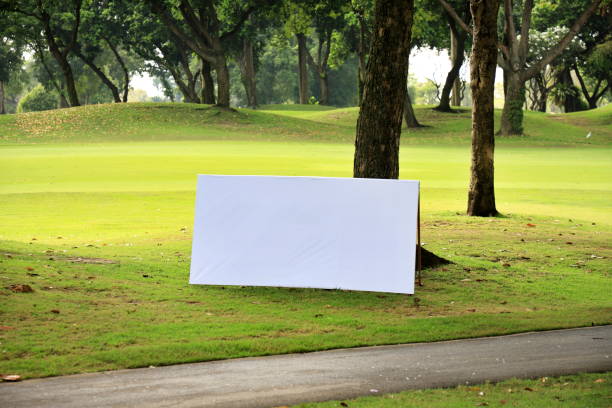 A blank white billboard sits on the grass at the base of a tree. The background is a blur of trees and grass on a golf course in Thailand in the daytime. In front there is a small cement road passing