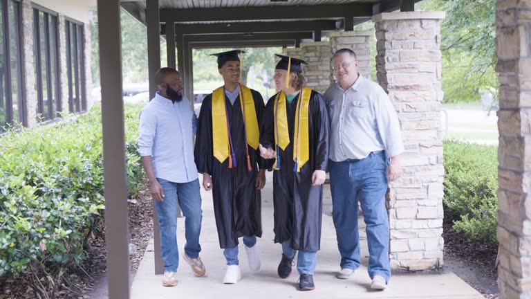 Two multiracial high school graduates walk with fathers