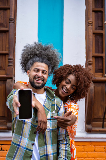 Young smiling latin couple between 25 - 30 years old showing empty iPhone screen on the street, the background wall has bright color tones located in the neighborhood la candelaria in the city of bogota ( colombia ).