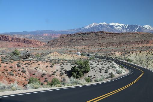 The Arches National Park in Moab Utah is renowned for its diverse collection of natural arches, towers, and other geological wonders, making it a paradise for outdoor enthusiasts and photographers alike. A camper travels along the park road and crosses through the desert with the La Sal Mountain range in the background.