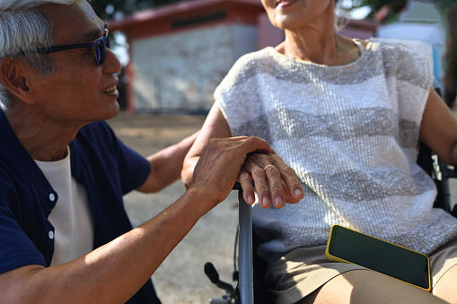 A caring elder man holding an elderly lady's hands, the old man holding a hand to encourage a senior woman in a wheelchair. Elderly care concept.