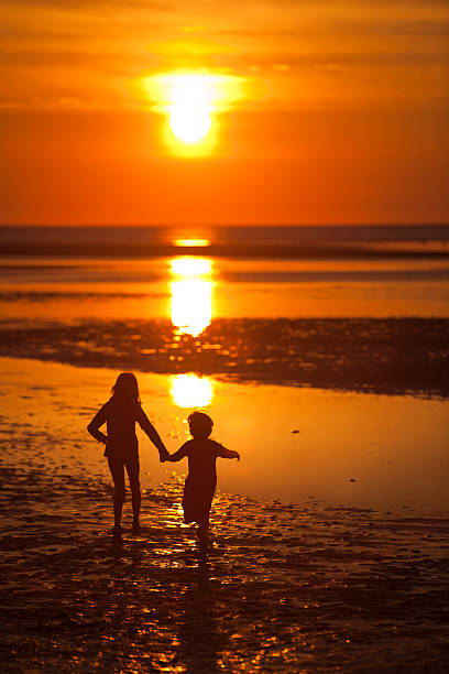 A little girl and boy hold hands as they walk into the sunset on a beach at low tide.