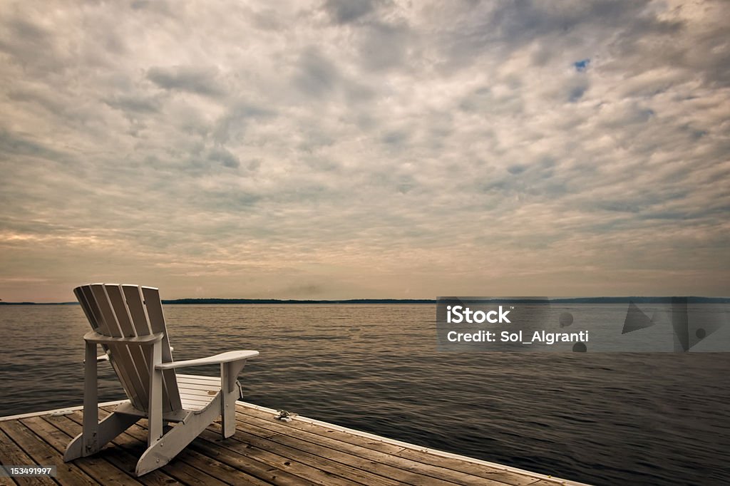 Tranquility This photo was taken in the summer late afternoon.  The vertical boards of the chair against the horizontal boards of the dock are creating a graphic composition.  In addition the white color of Muskoka chair is complementing the white sky. Backgrounds Stock Photo