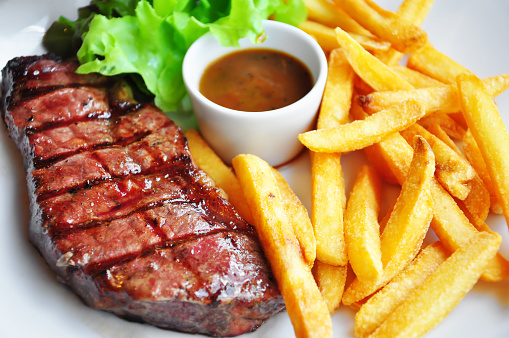 A plate of grilled beef steak, lettuce, French fries and a container of sauce at the center.  The steak is oozing juice, the lettuce is fresh, the sauce is brown, and the fries are golden.