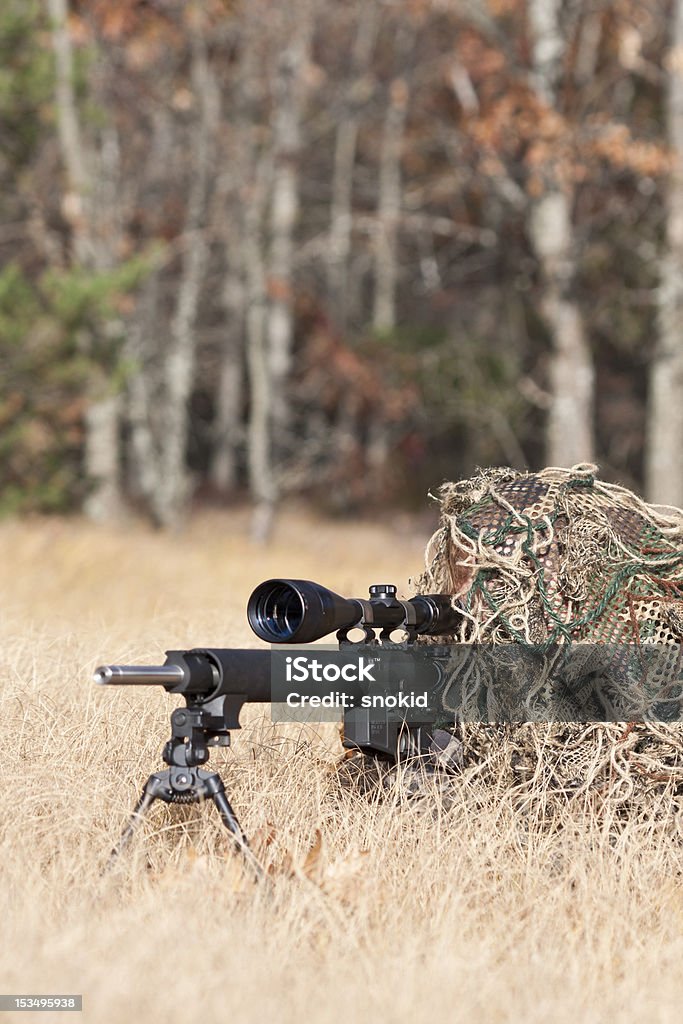 sniper sniper laying on the ground covered in a ghille suite tall grass and trees in the background Addiction Stock Photo