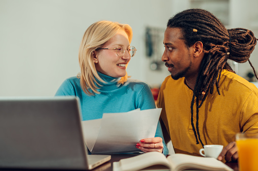 A cheerful interracial couple is doing home finances on the laptop. A happy woman is holding paperwork and bills while smiling at her Arabic husband. The spouses are paying bills and taxes from home.