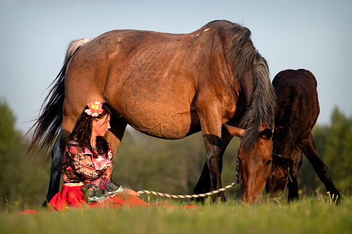 A gypsy-dressed girl sits near her horses in the field at sunset.