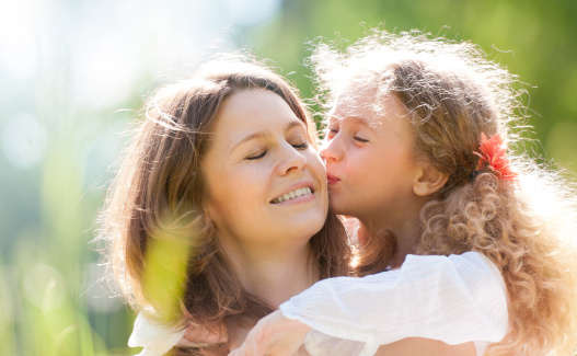 Little daughter kissing her young beautiful mother, their eyes closed, both smiling. Nature background.