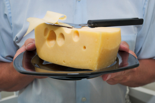 A man holding a large cheese on a plate, with a cheese slicer on top.