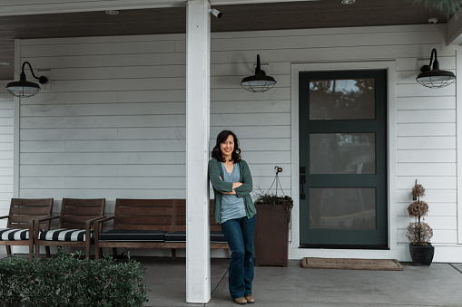 Portrait of a Vietnamese woman smiling confidently at the camera while leaning casually against a pillar on the covered front patio of her modern farmhouse style home.