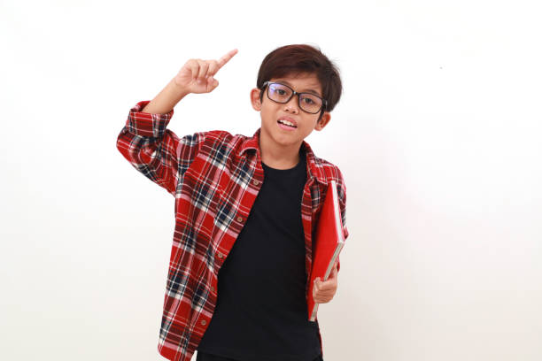 Happy smart asian boy holding a book while raising his hand. Isolated on white