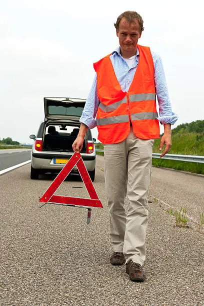 Man walking backwards from his car wearing a safety vest, carrying a warning triangle