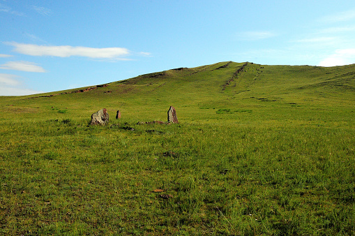 Ancient stone menhirs in a wide clearing overgrown with tall grass at the foot of a high mountain with parallel stone formations. Mount Uitag, Khakassia, Siberia, Russia.
