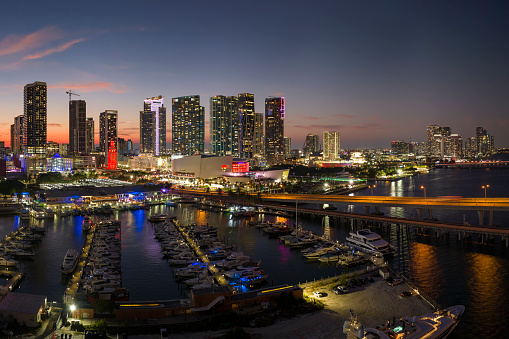 Aerial view of yachts in Miami marina at Bayside Marketplace with reflections in Biscayne Bay water and high skyscrapers of Brickell, city's financial center at night.