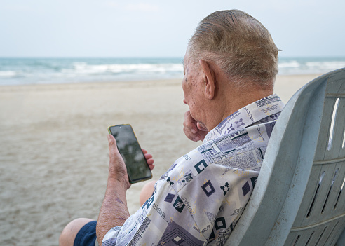 Senior man sitting on a beach chair and using his smartphone to read. Using technology for the elderly.