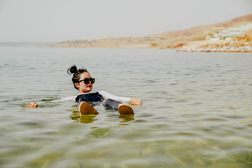 An Asian tourist traveler floating in the therapeutic water of the dead sea, placed in border of Israel and Jordan