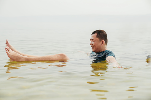 An Asian tourist traveler floating in the therapeutic water of the dead sea, placed in border of Israel and Jordan