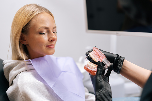 Close-up cropped shot of unrecognizable female dentist in protective mask and gloves showing jaw model with braces to young woman patient in dental office. Concept of stomatology, orthodontics.