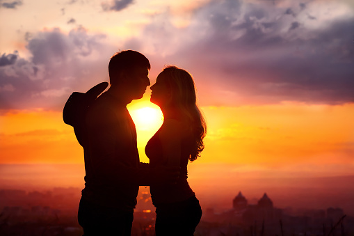 Young couple silhouette hugging and kissing outdoors at sunset background. Sun between them. Man with cowboy hat at his back