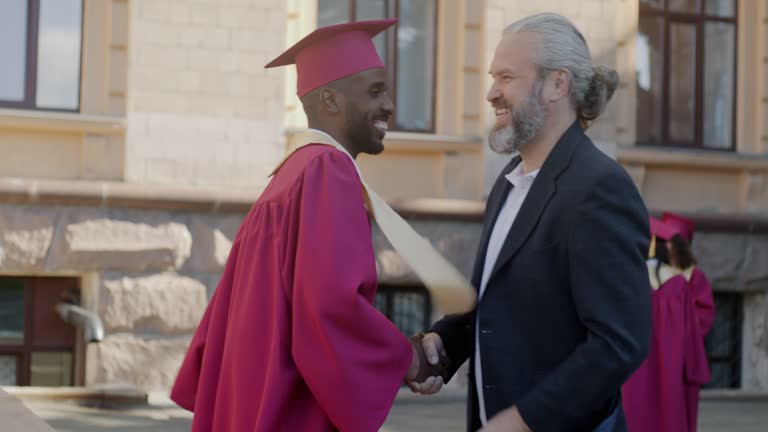 College teacher talking to graduate student in mortar board and gown shaking hands and hugging man on campus