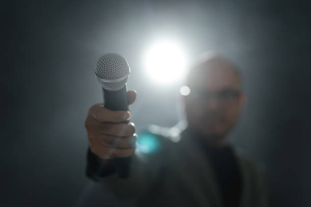 istock Confident successful speaker man talking on stage with spotlight strike through the darkness at corporate business event. Public speaker giving talk at conference hall. Stand up comedian. 1535648484