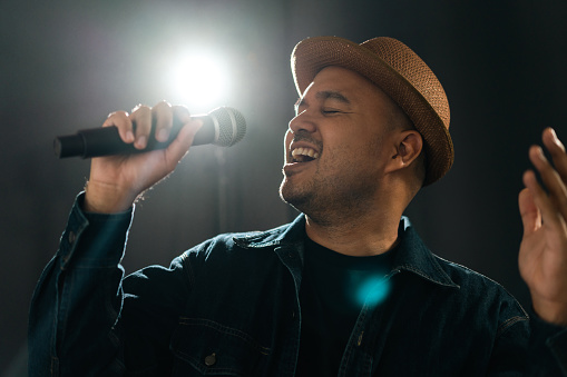 Close up shots of Attractive singer man singing music and playing guitar on stage with spotlight strike through the darkness at concert event. Musician performing live with light and smoke at hall.