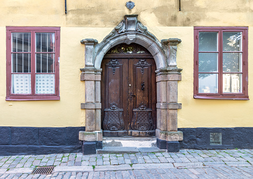 Vintage decorated wooden door framed by stone engraved frame, mediating three wooden and wrought iron windows in a grunge yellow wall at street with cobblestone floor, Old town, Stockholm, Sweden