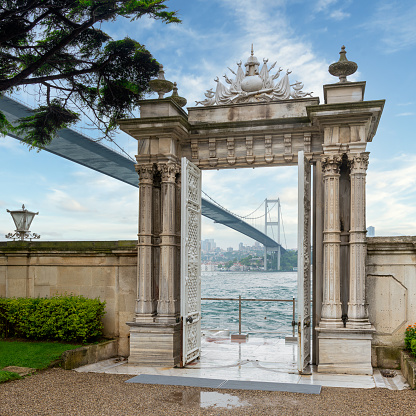 Marble gate with decorated open door leading to the Bosphorus Strait, framing Bosphorus Bridge, located at Beylerbeyi Palace, Istanbul, Turkey. Text above translate: In the name of God