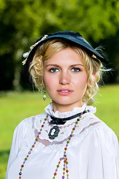 A headshot of an attractive young victorian era woman. She is bedecked in jewelry and a hat with a frilly white top