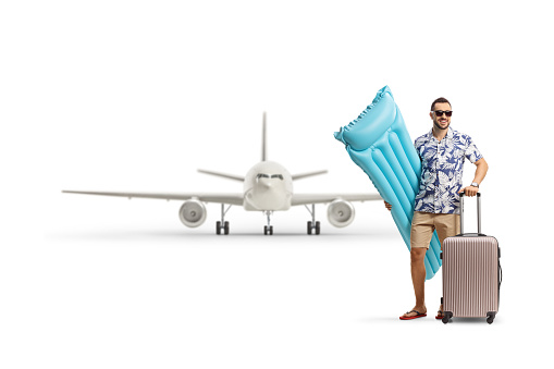 Full length portrait of a man holding an inflatable mattress and a suitcase in front of a plane isolated on white background