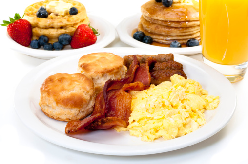Breakfast plate with scrambled eggs, bacon, and buttermilk biscuits.  Waffles, pancakes, and orange juice in background.  Isolated on white background.